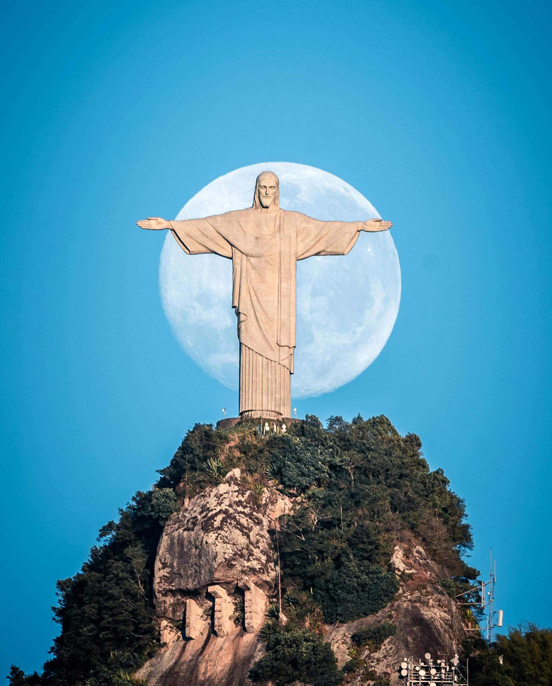 Cristo Redentor no Rio de Janeiro visto de frente, com a lua cheia ao fundo e céu azul